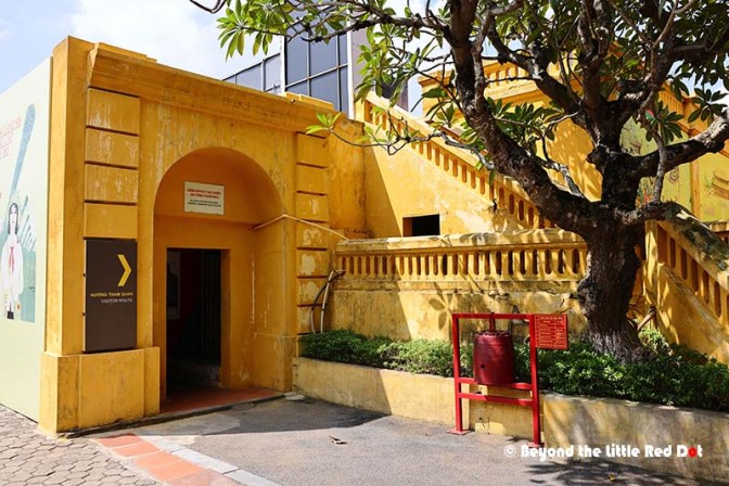 A yellow building entrance with a curved archway leading to a staircase, greenery around, and a red fire hydrant in the foreground.