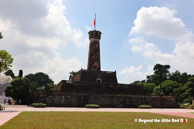A tall historical tower with a flag at the top, surrounded by green trees and a clear blue sky, located in a landscaped area.