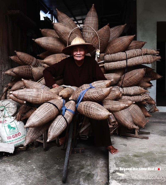 An elderly man smiling while sitting in front of a large arrangement of woven bamboo baskets, resembling a flower shape, displaying a traditional crafting technique.