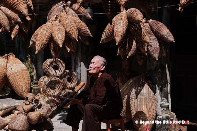 An elderly man sitting contently among various woven baskets and fishing nets hung above him in a traditional workshop.