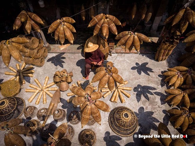 Aerial view of a traditional artisan working among hanging woven baskets and decorative items made of bamboo, showcasing intricate craftsmanship in a well-lit outdoor setting.