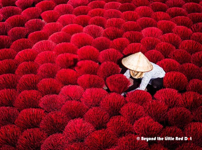 A person wearing a conical hat kneeling amidst a large field of vibrant red incense sticks arranged closely together.