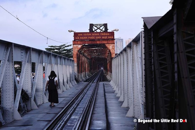 A woman walking along a railway bridge with metal supports and a sign overhead, under a cloudy sky.