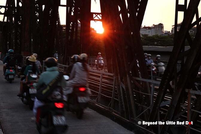 Motorcycles crossing a bridge during sunset, with city buildings in the background.