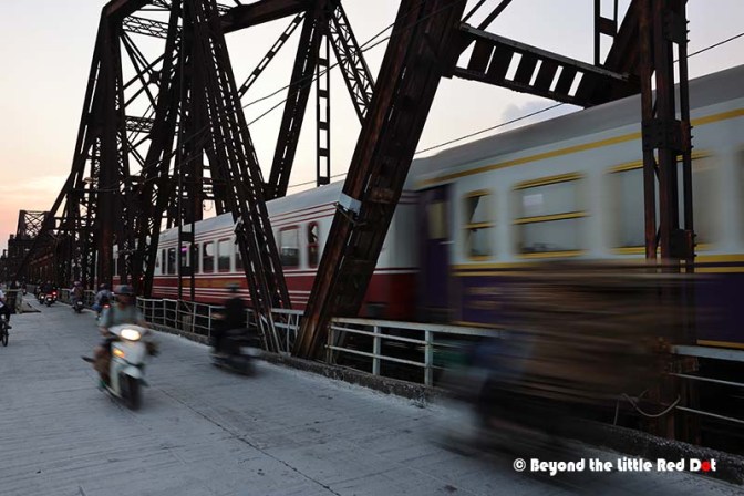 A motion blur image of a train crossing a bridge at sunset, with motorcycles riding on the adjacent pathway.