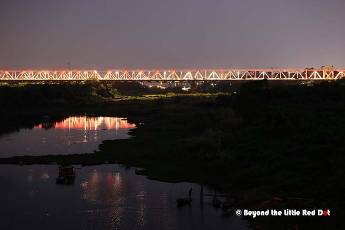 Night view of a bridge illuminated against a dark sky, reflecting light on a nearby river with boats in the foreground and lush vegetation along the banks.
