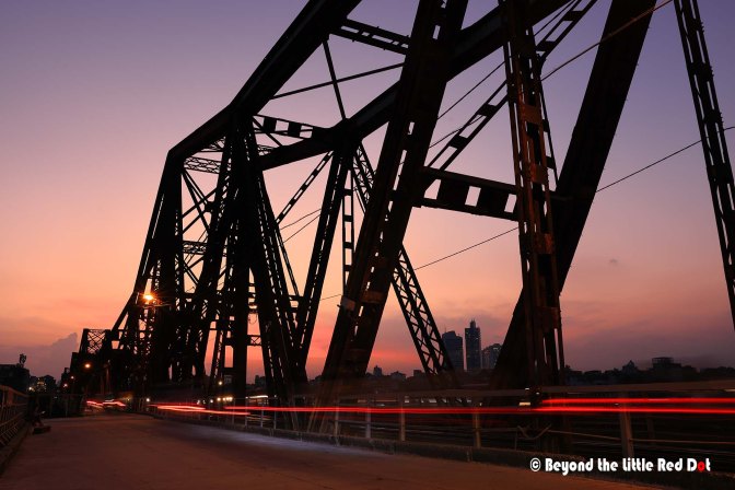 Silhouette of a metal bridge against a colorful sunset sky, with city skyline in the background and light trails from vehicles on the road.