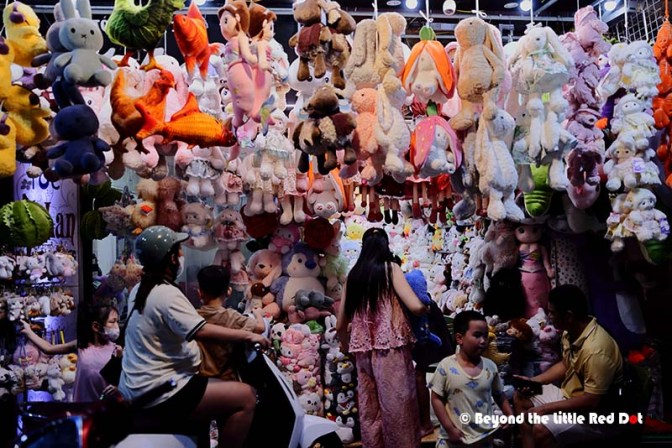 A vibrant scene at a toy store filled with various plush toys and stuffed animals hanging from the ceiling, with shoppers including children and adults browsing among them.