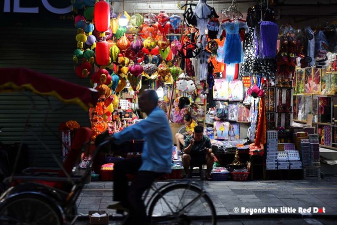 A vibrant street market at night, showcasing colorful lanterns and festive decorations. A person rides a bicycle in the foreground, while two others sit in front of a stall filled with various merchandise.