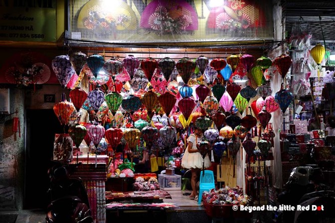 A colorful shop filled with hanging decorative lanterns in various shapes and patterns, illuminated at night. A person is partially visible in the shop, surrounded by other decorative items.