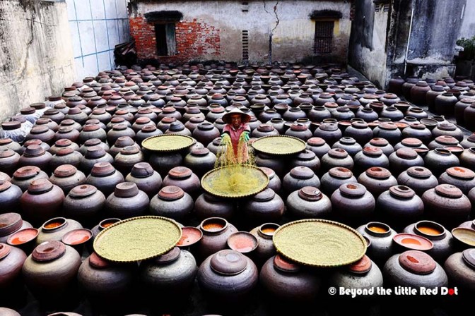 A traditional ceramic pottery workshop with numerous clay jars arranged in rows, while a person in a straw hat is handling grains in the center.