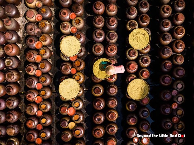 Aerial view of numerous small clay pots arranged in rows, with some topped by gold-colored lids, featuring a pink stopper on one of the pots.