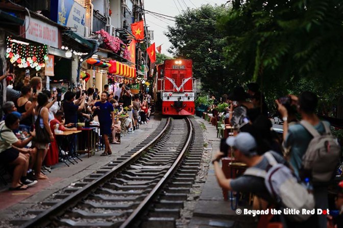 A vibrant street scene in Vietnam featuring a train approaching amidst a crowd of people sitting at outdoor tables along the railway track, adorned with colorful flags and flowers.