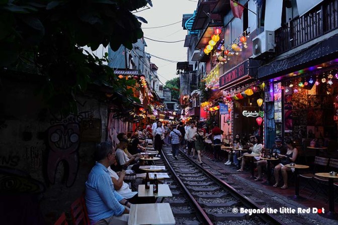 A vibrant street scene featuring a narrow alley lined with colorful lanterns, restaurants, and small tables. People are dining and socializing along the railway tracks, with buildings adorned with vibrant street art.